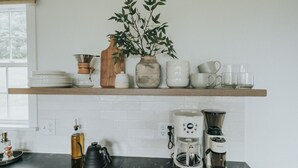 Une cuisine avec une étagère en bois où sont posées des assiettes, des tasses et une plante. En dessous, sur le plan de travail, on trouve des machines à café et une bouilloire.