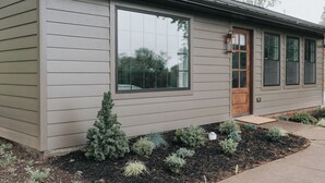 Une maison moderne dotée d’une grande fenêtre, d’une porte en bois et d’un petit jardin où poussent différentes espèces de plantes.