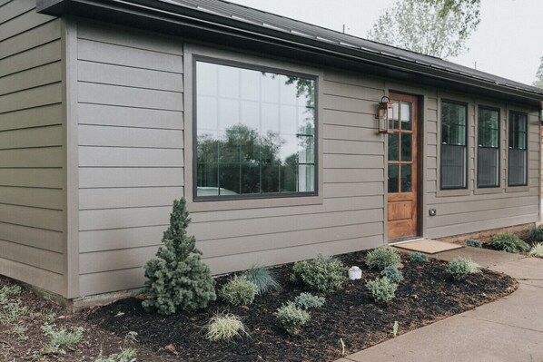 A modern house with a large window, a wooden door, and a small garden with various plants.