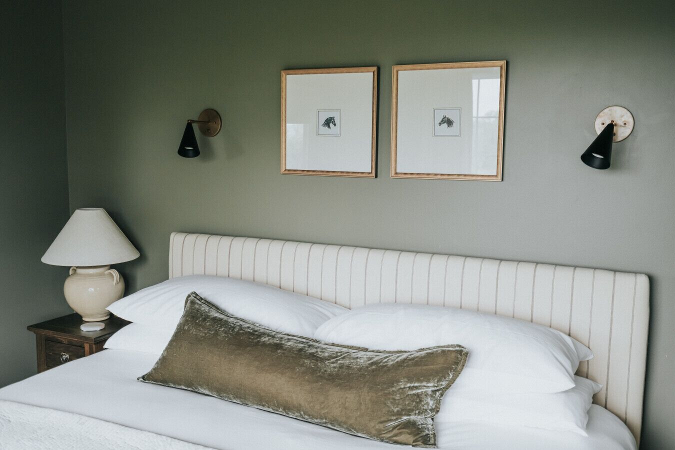 En pænt redt seng med hvide sengetøj og en brun pude, et natbord med en lampe, og to billeder hængende på væggen.