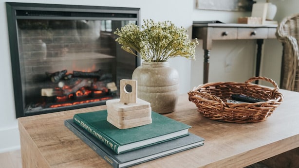 Una mesa de madera con un libro verde, un bloque de madera pequeño, un florero con flores y una cesta de mimbre.