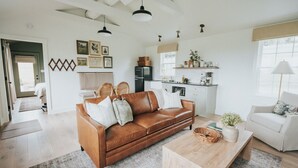 Amplia sala de estar con un sofá de cuero marrón, una mesa de centro de madera y un sillón blanco. Se observa una zona de cocina con armarios blancos y un refrigerador.