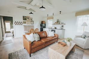 Amplia sala de estar con un sofá de cuero marrón, una mesa de centro de madera y un sillón blanco. Se observa una zona de cocina con armarios blancos y un refrigerador.