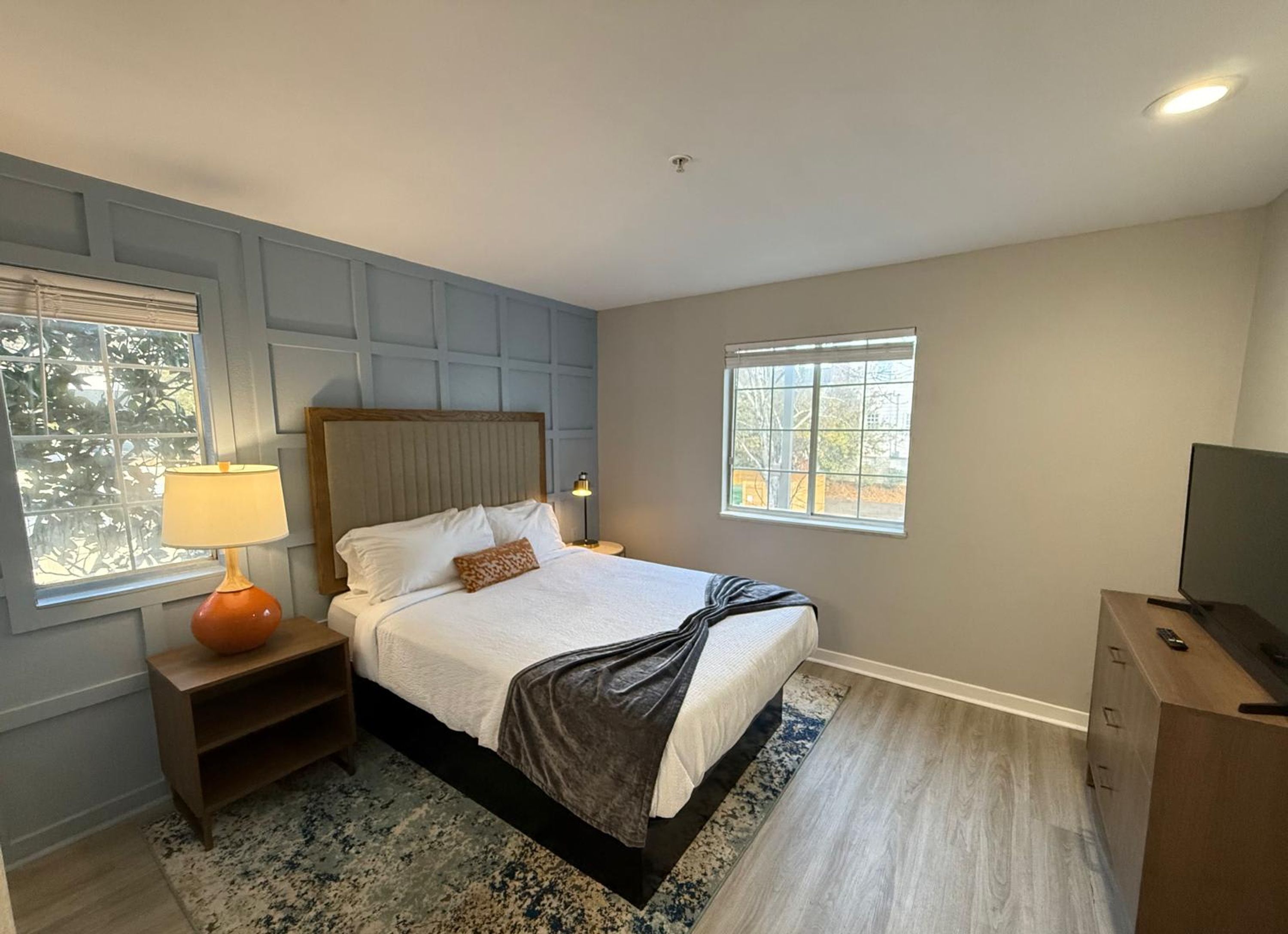 A bedroom with a bed, nightstands, a lamp, a TV, and two windows.