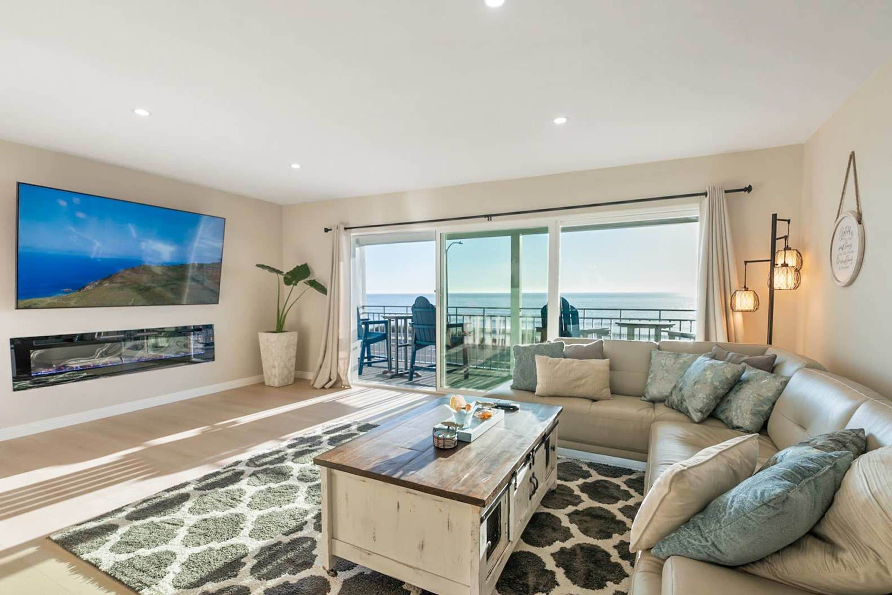 A living room with a sofa, a coffee table, a TV mounted on the wall, and a large window with a view of the sea.