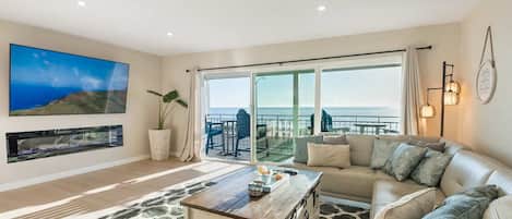 Un salon comprenant un canapé, une table basse, un téléviseur fixé au mur et une grande fenêtre donnant sur la mer.