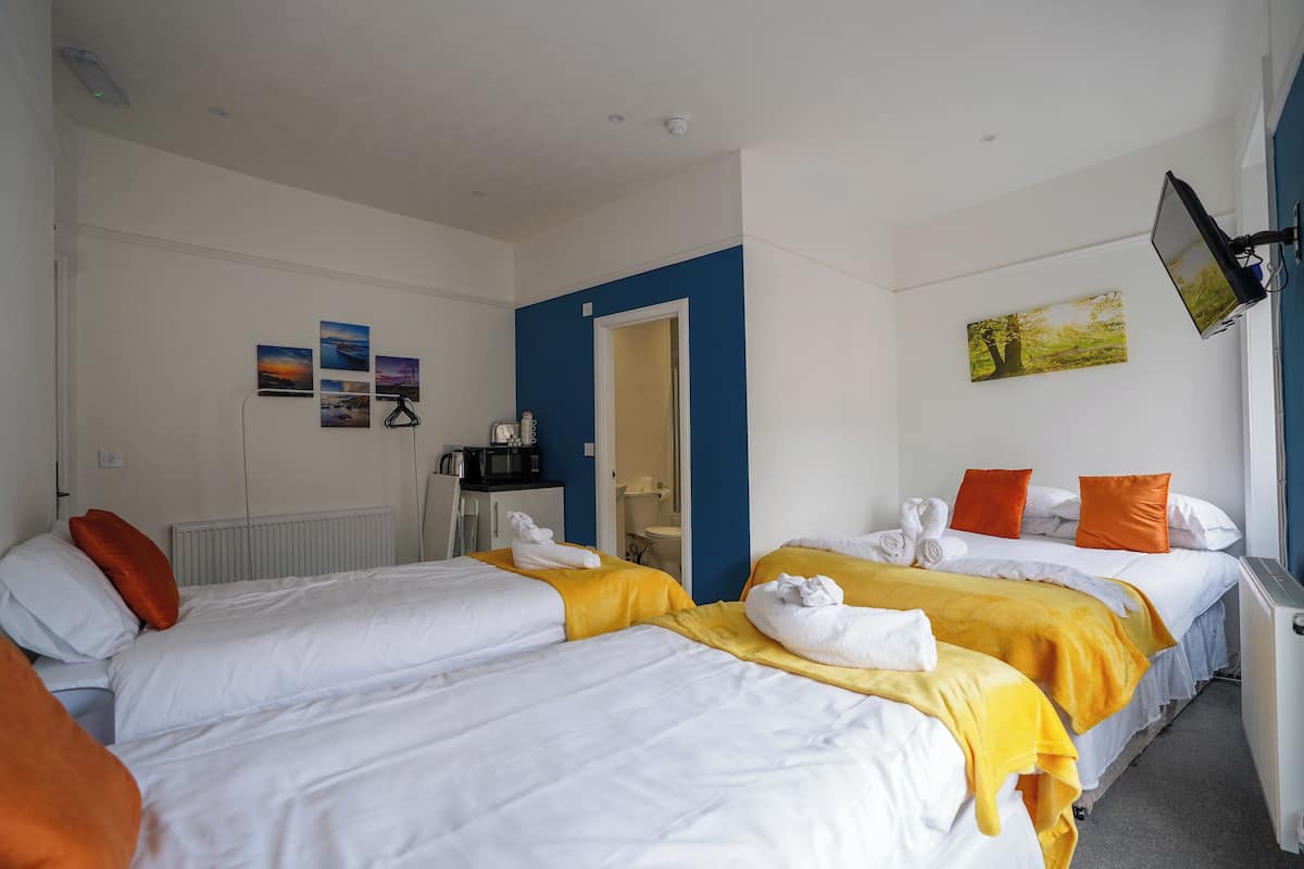 Habitación de hotel con dos camas, cada una con sábanas blancas y almohadas naranjas. Hay toallas sobre las camas, una mesita con una tetera y un cuadro en la pared.