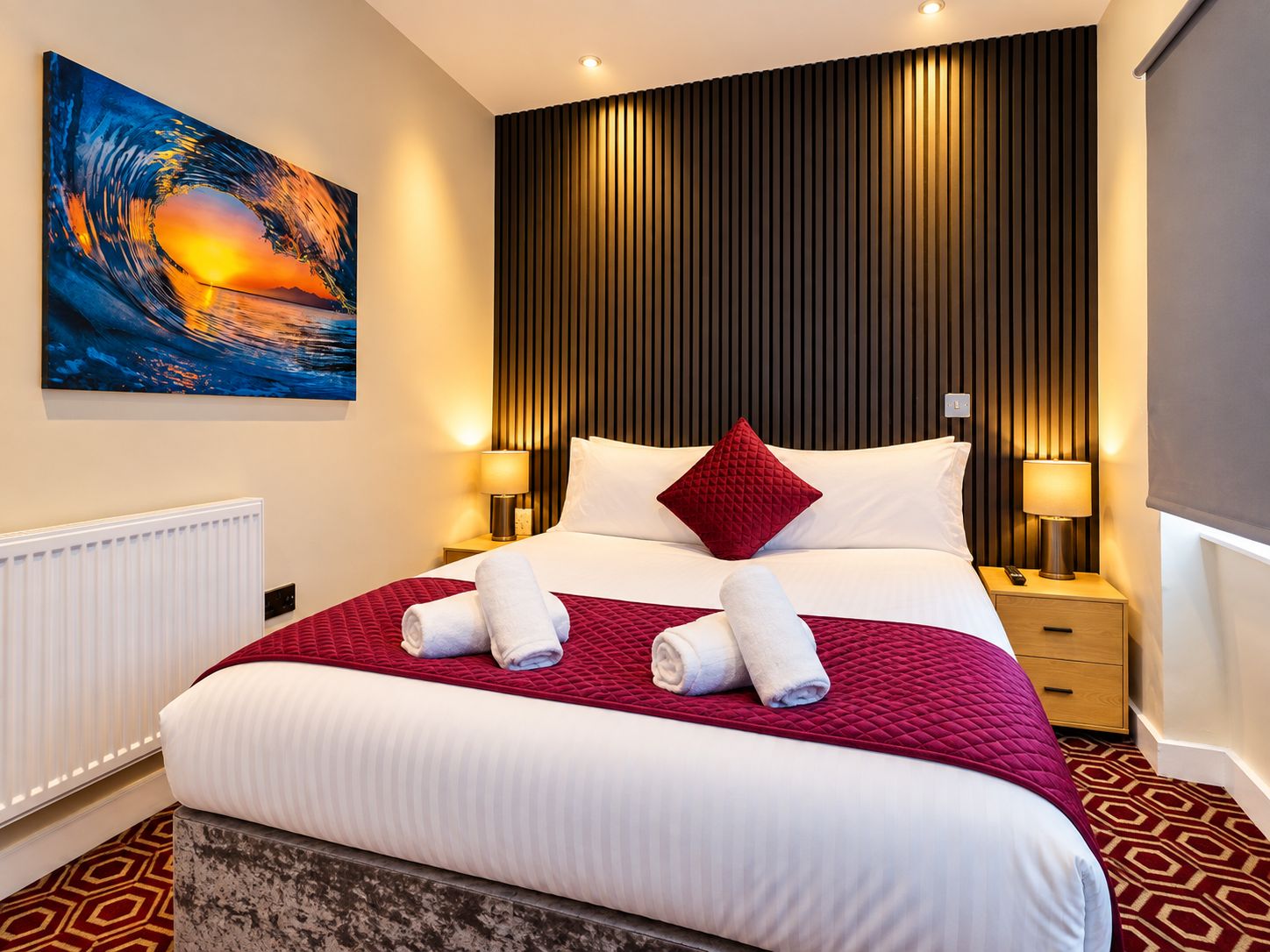 A hotel room with a large bed, a red quilted bedspread, white linens, a red pillow, two bedside lamps, and a painting of a sunset on the wall.