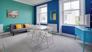 Une pièce aux murs de couleur turquoise, avec un canapé gris agrémenté de coussins jaunes, une table blanche avec des chaises, une commode bleue et une fenêtre donnant sur des immeubles.