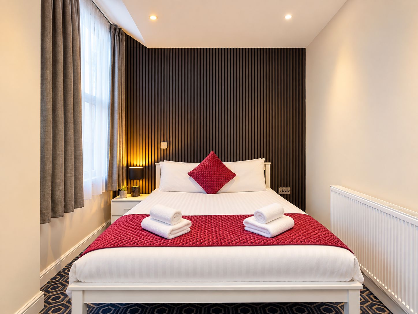 Una habitación de hotel con cama, mesita de noche, lámpara y ventana con cortinas.
