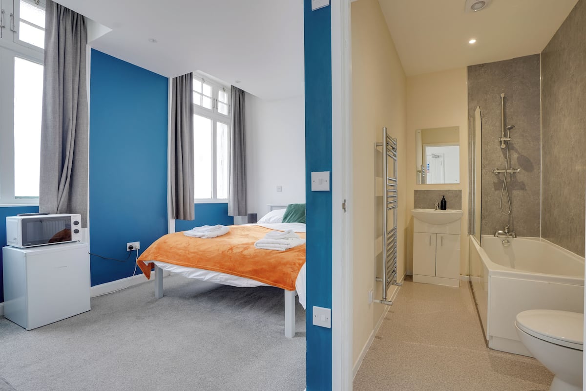 Un dormitorio con una cama, microondas y ventana con cortinas. Las paredes son azules y el techo, blanco.