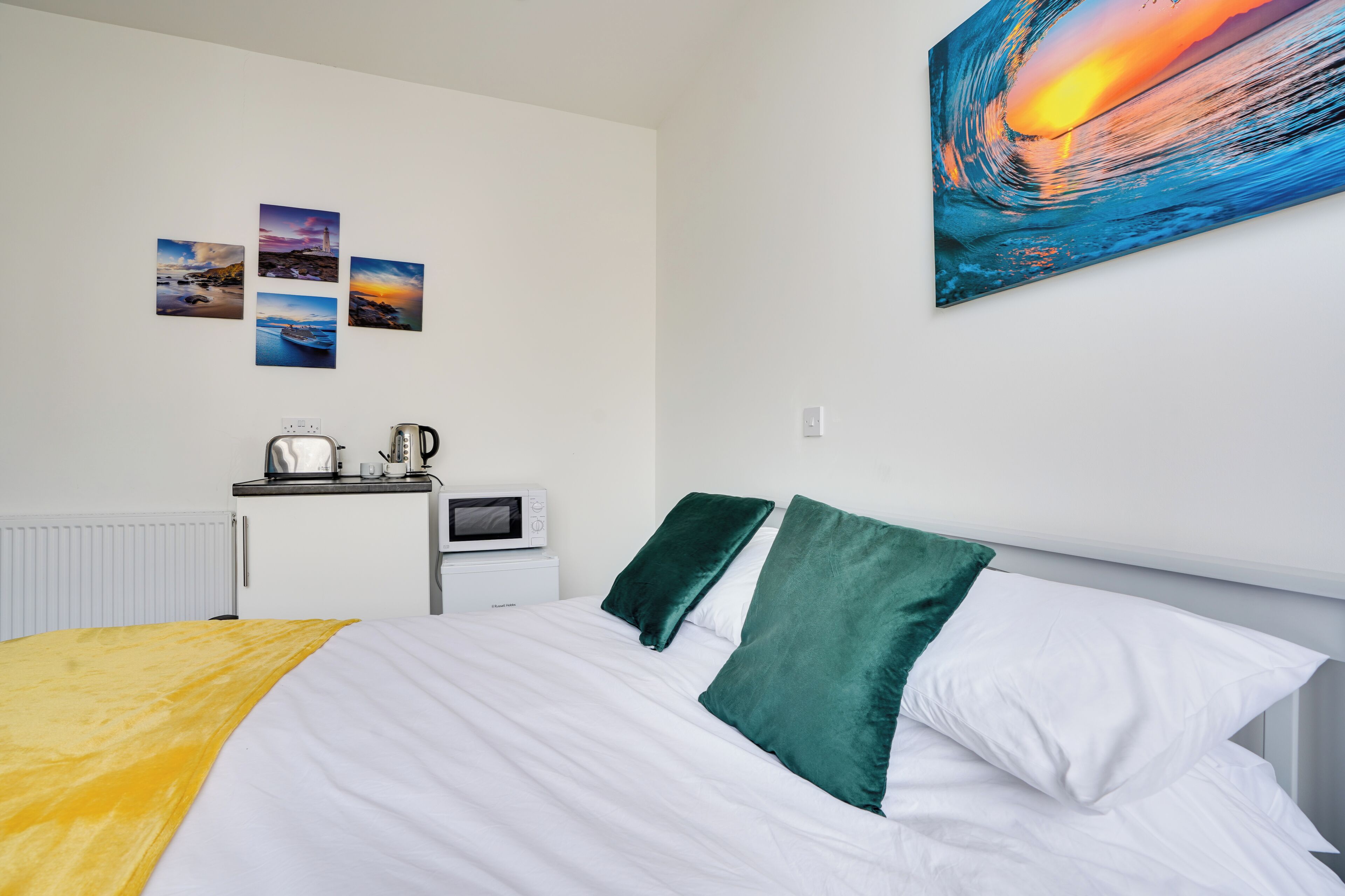 Una cama bien hecha con almohadas blancas y verdes, una colcha amarilla y una pared con cuatro cuadros enmarcados.