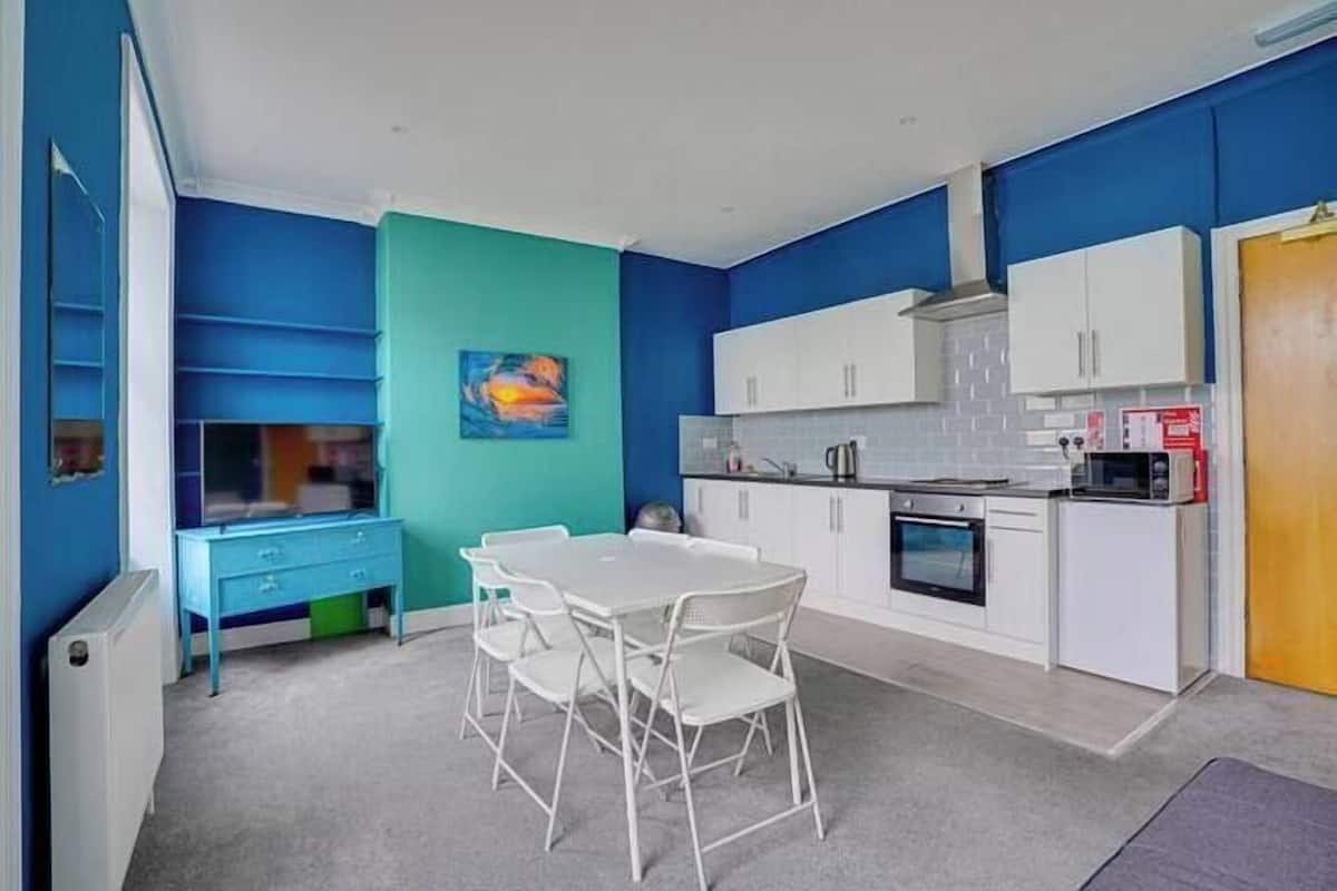 Una cocina compacta con armarios blancos, una pequeña mesa de comedor con sillas y una pared de acento azul.