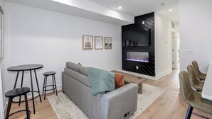 A modern living room with a grey sofa, black bar stools, a flat-screen TV, and a fireplace. - Capitol Hill Suites - Unit 9 (Washington)