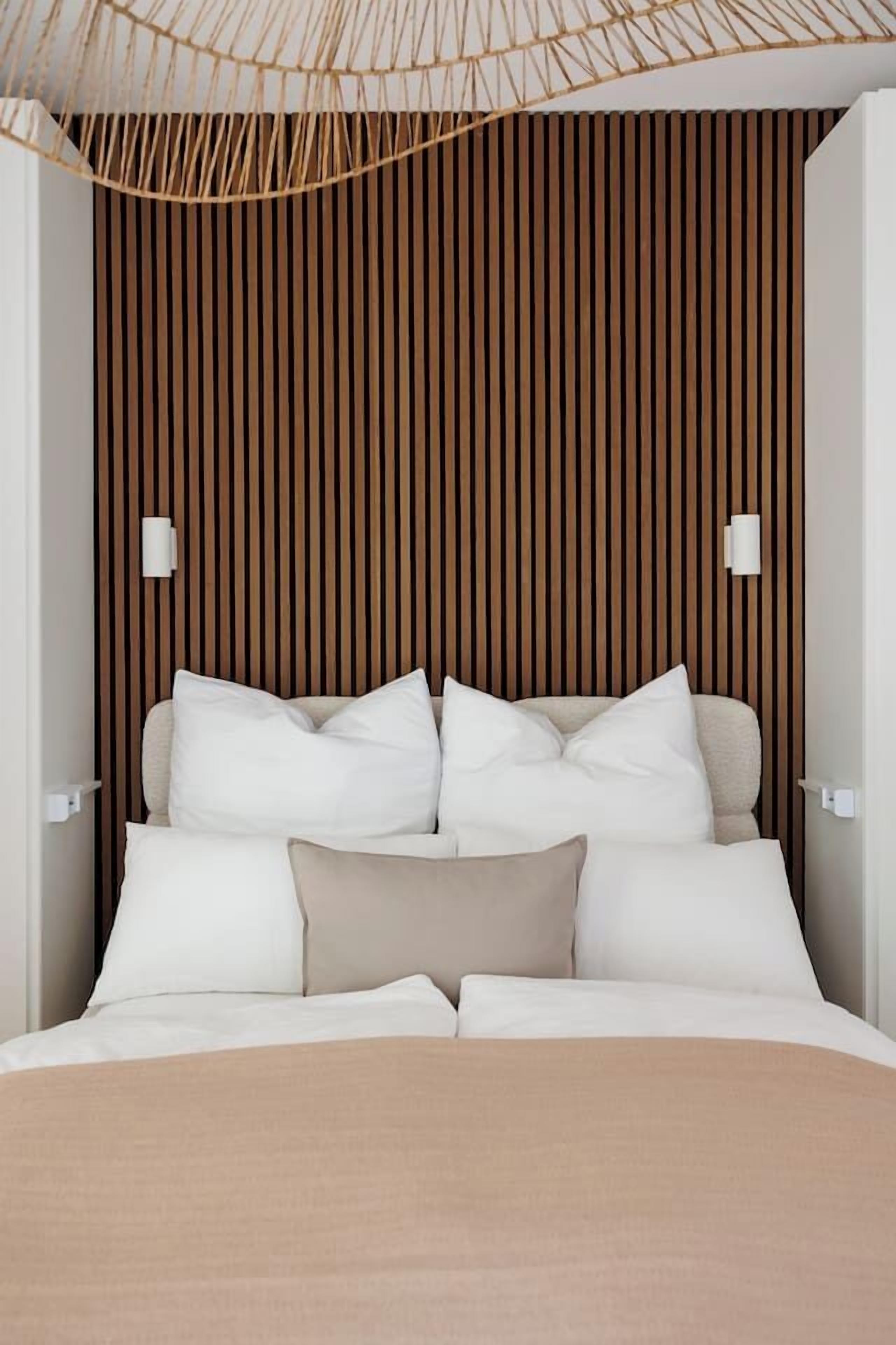 Ein Bett mit weißer und beiger Bettwäsche, eine Wand aus Holzleisten und zwei an der Wand befestigte Leuchten.