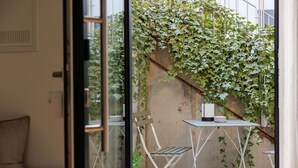 A room with a glass door leading to a balcony with a white table and chairs, a candle, and ivy-covered walls.
