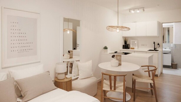 A modern, minimalist living room with a sofa, a round dining table, and a kitchen area with white cabinets and a hanging light fixture.