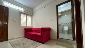 A room with a red sofa, a wooden door, a window with blinds, and a bathroom visible through a glass door. - Hsr Lodestone By Newstays (BANGALORE)