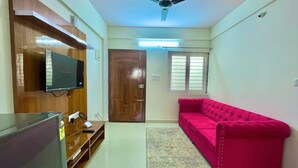 A living room with a flat-screen TV mounted on a wooden cabinet, a ceiling fan, a wooden door, a window with blinds, and a bright red sofa. - Hsr Lodestone By Newstays (BANGALORE)