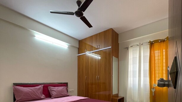 A bedroom with a bed, a ceiling fan, a wardrobe, and a window with curtains. - Hsr Lodestone By Newstays (BANGALORE)