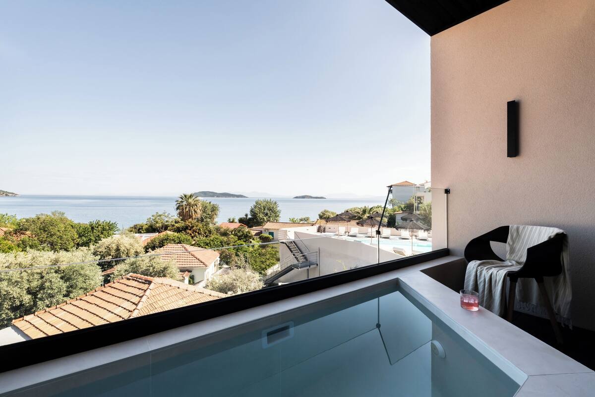 A modern balcony with a glass barrier, a pool, and a view of the sea and surrounding buildings.