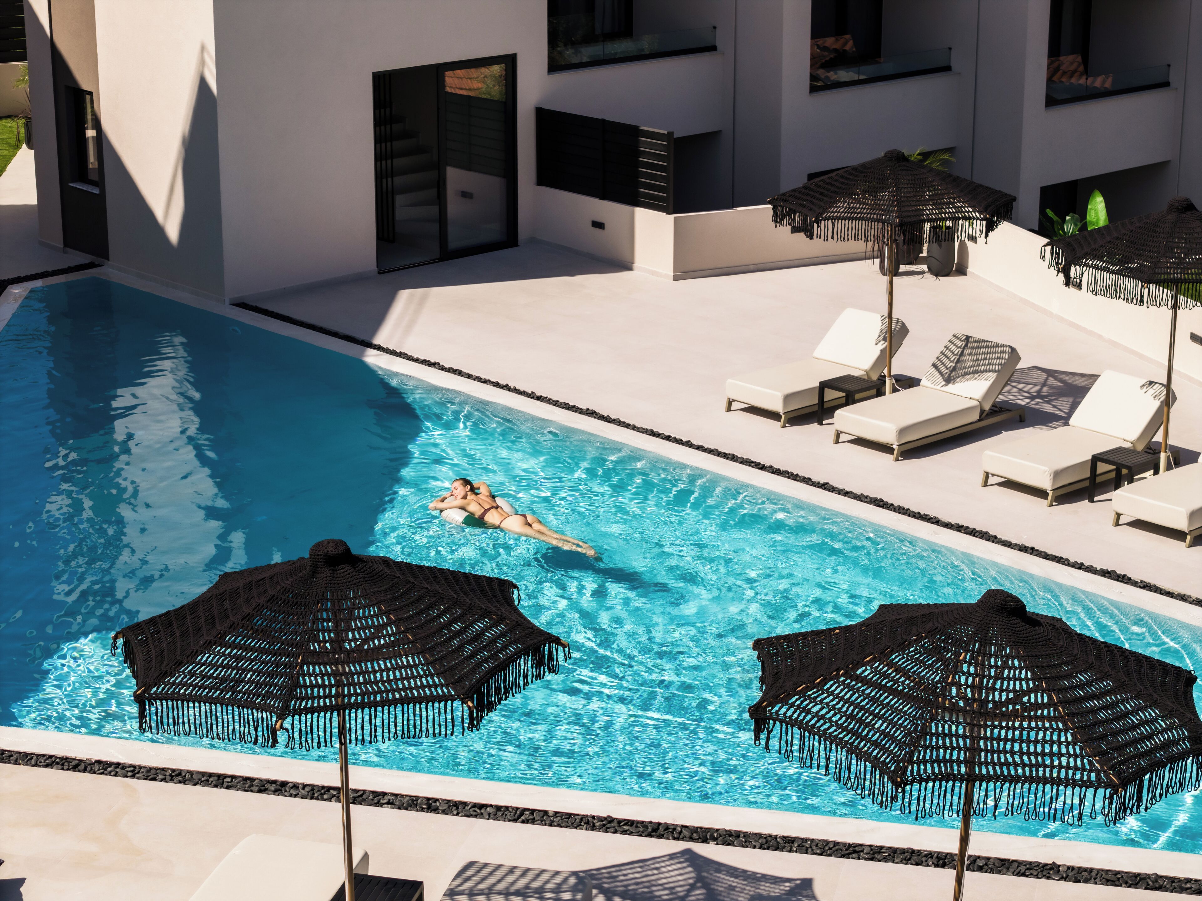 Una piscina con sillones de descanso y sombrillas, rodeada por un edificio moderno con grandes ventanales.