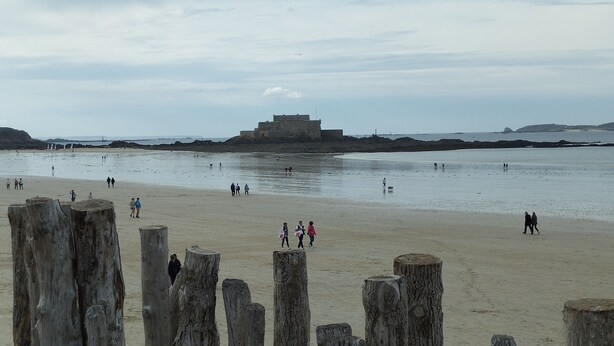 Ein Strand mit Holzbalken im Vordergrund, einem sandigen Ufer und einem großen Gebäude im Hintergrund.