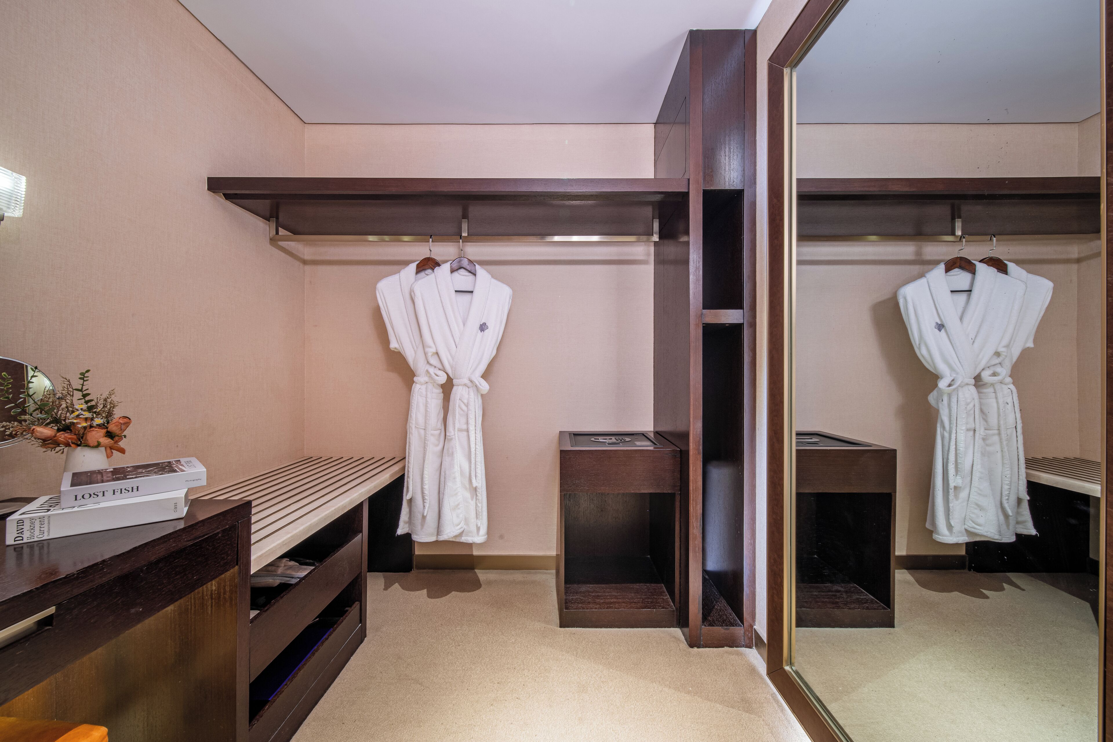 Ett hotellbadrum med två vita badrockar hängande på träkrokar, en bänk med en bok som heter ”FÖRLORAD FISK” på, och en spegel som reflekterar rummet.