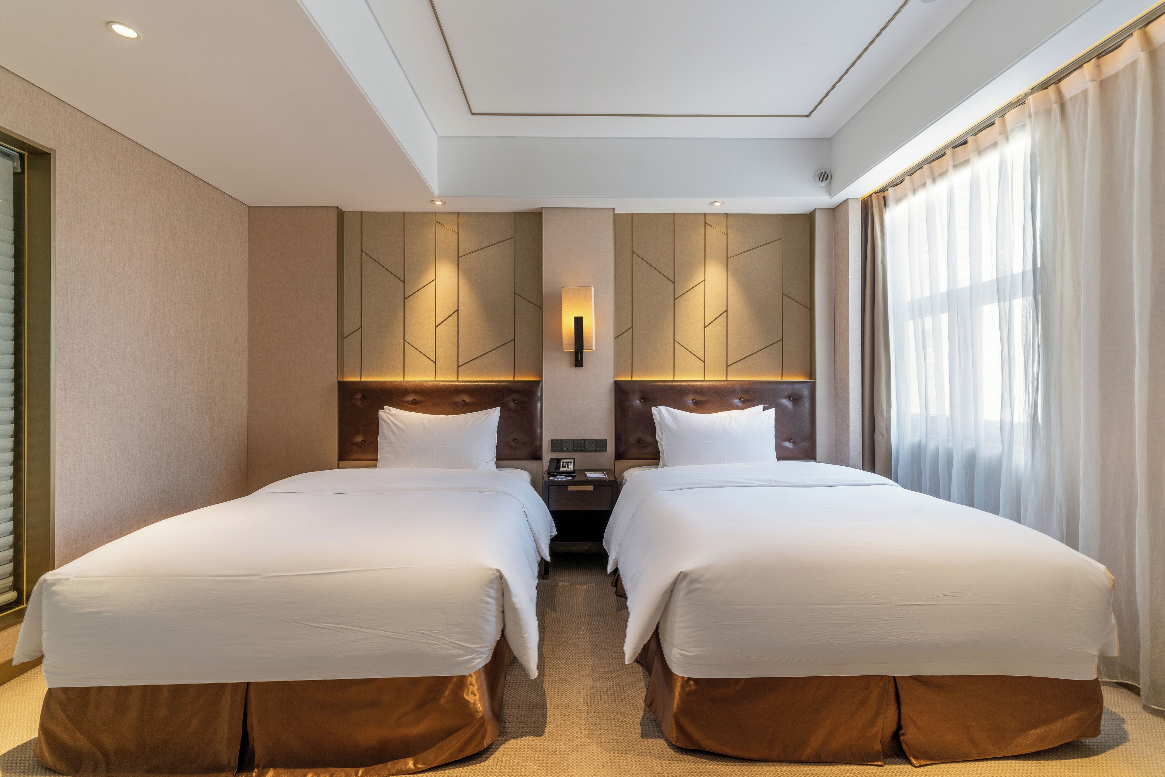 A hotel room with two beds, a nightstand, a wall-mounted light, and a large window with curtains.