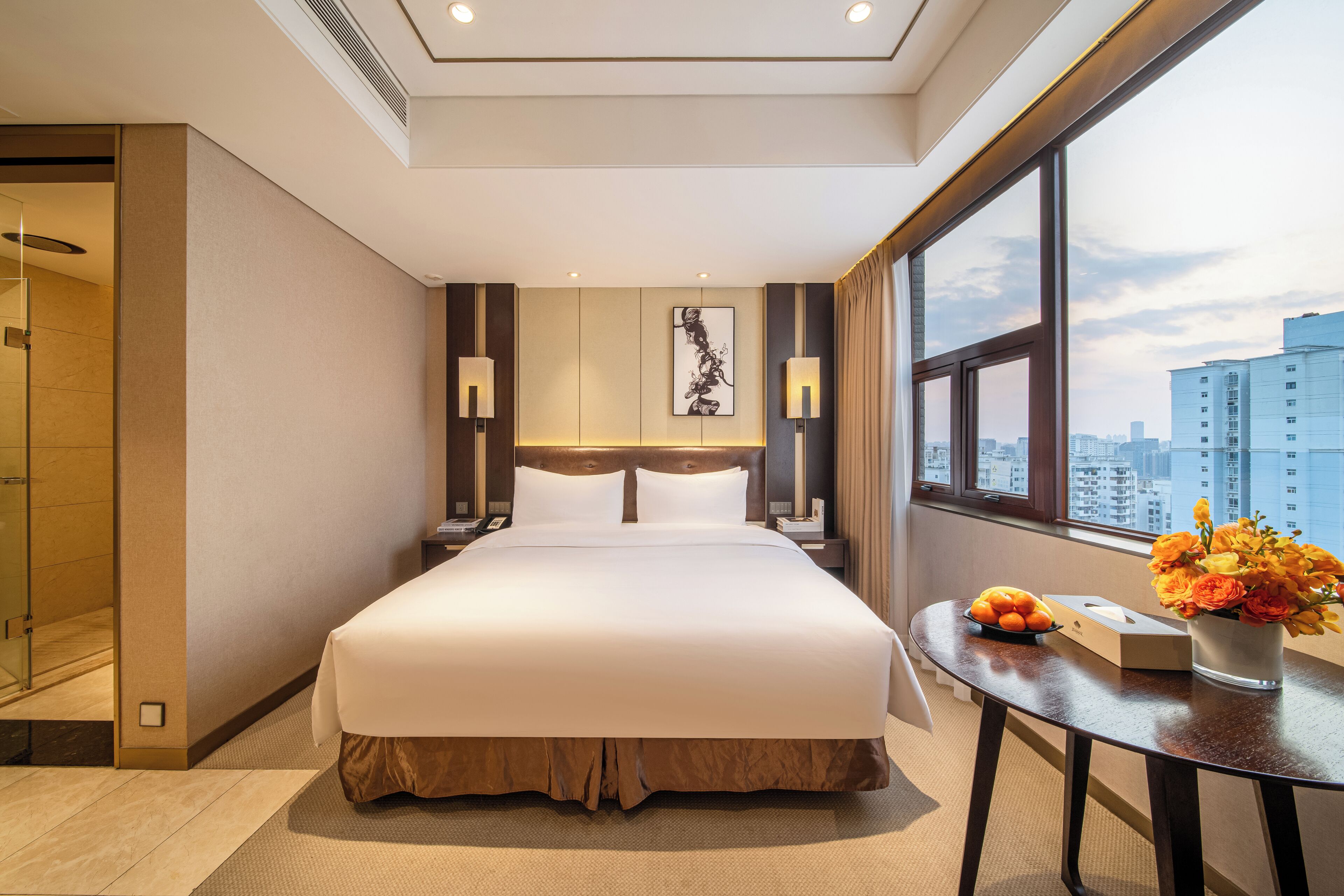 Habitación de hotel con una cama grande, una mesita de noche con un jarrón de flores y vistas a la ciudad a través de la ventana, con un diseño moderno.