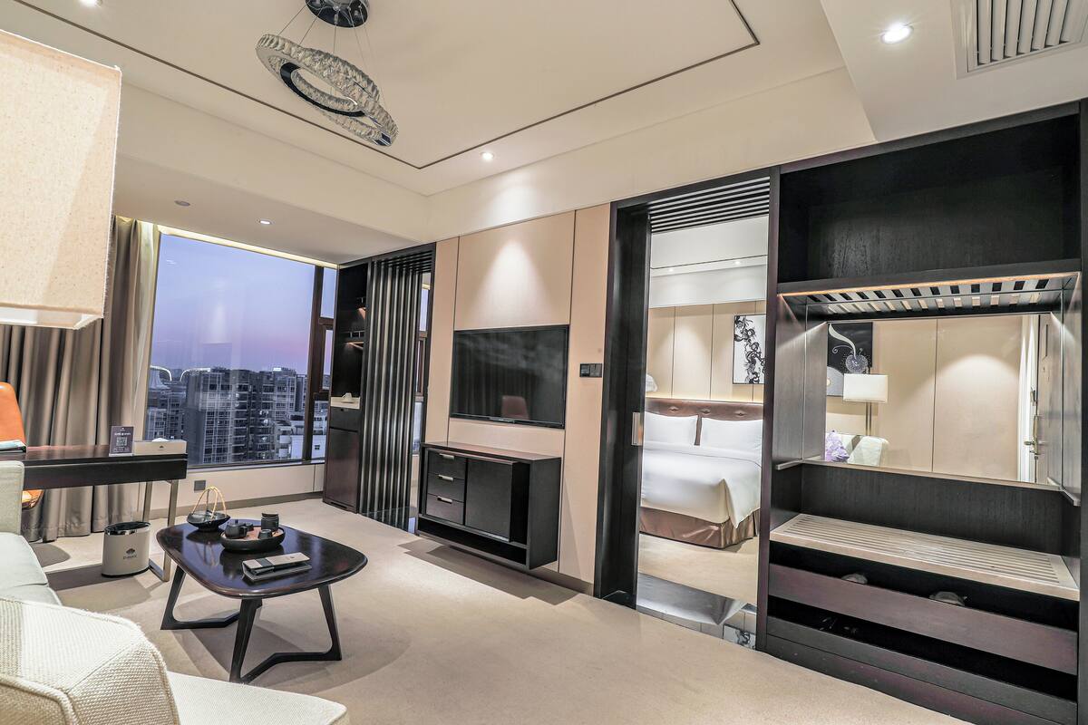A modern hotel room with a large window, a flat-screen TV, a sofa, a coffee table, and a built-in wardrobe.