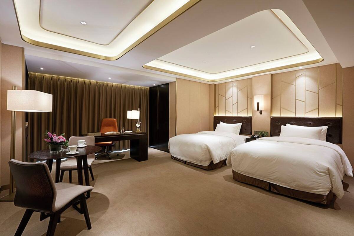 A hotel room with two beds, a desk with a chair, a lamp, and a dining table with chairs.