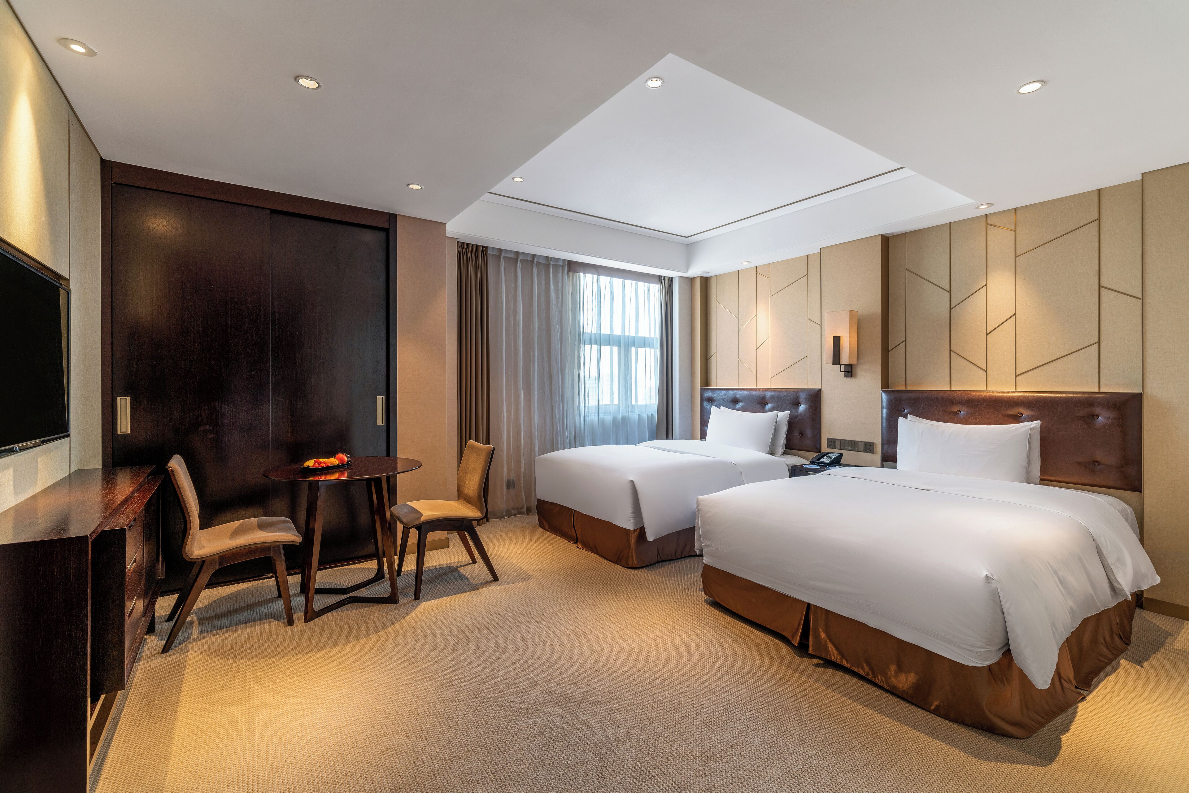 A hotel room with two beds, a desk, a chair, a TV, and a large window with curtains.