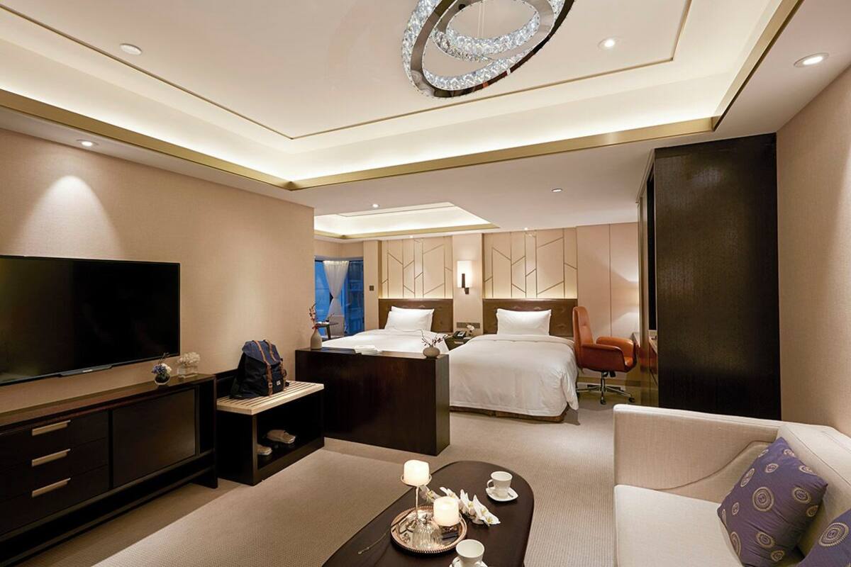 A modern hotel room with a large flat-screen TV, a bed with white linens, a sofa, a coffee table with a tray, and a decorative ceiling light.