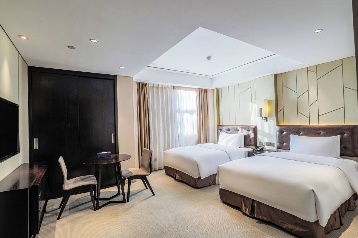 A hotel room with two beds, a desk, a chair, a TV, and a large window with curtains.
