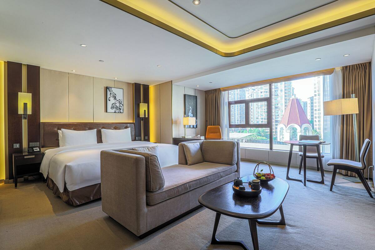 A modern hotel room with a bed, sofa, coffee table, and a view of the cityscape.