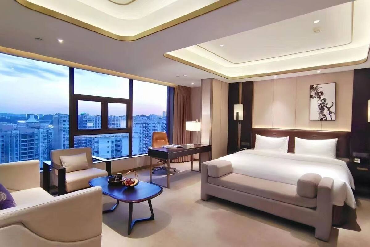 A modern hotel room with a large bed, a sofa, a desk, and a view of the cityscape.