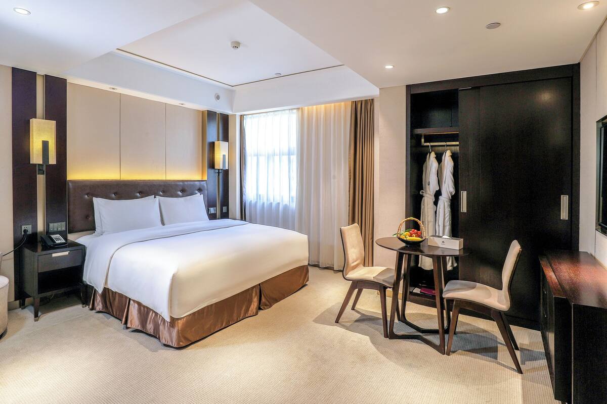 A hotel room with a large bed, bedside table, lamp, and a small table with a fruit bowl.