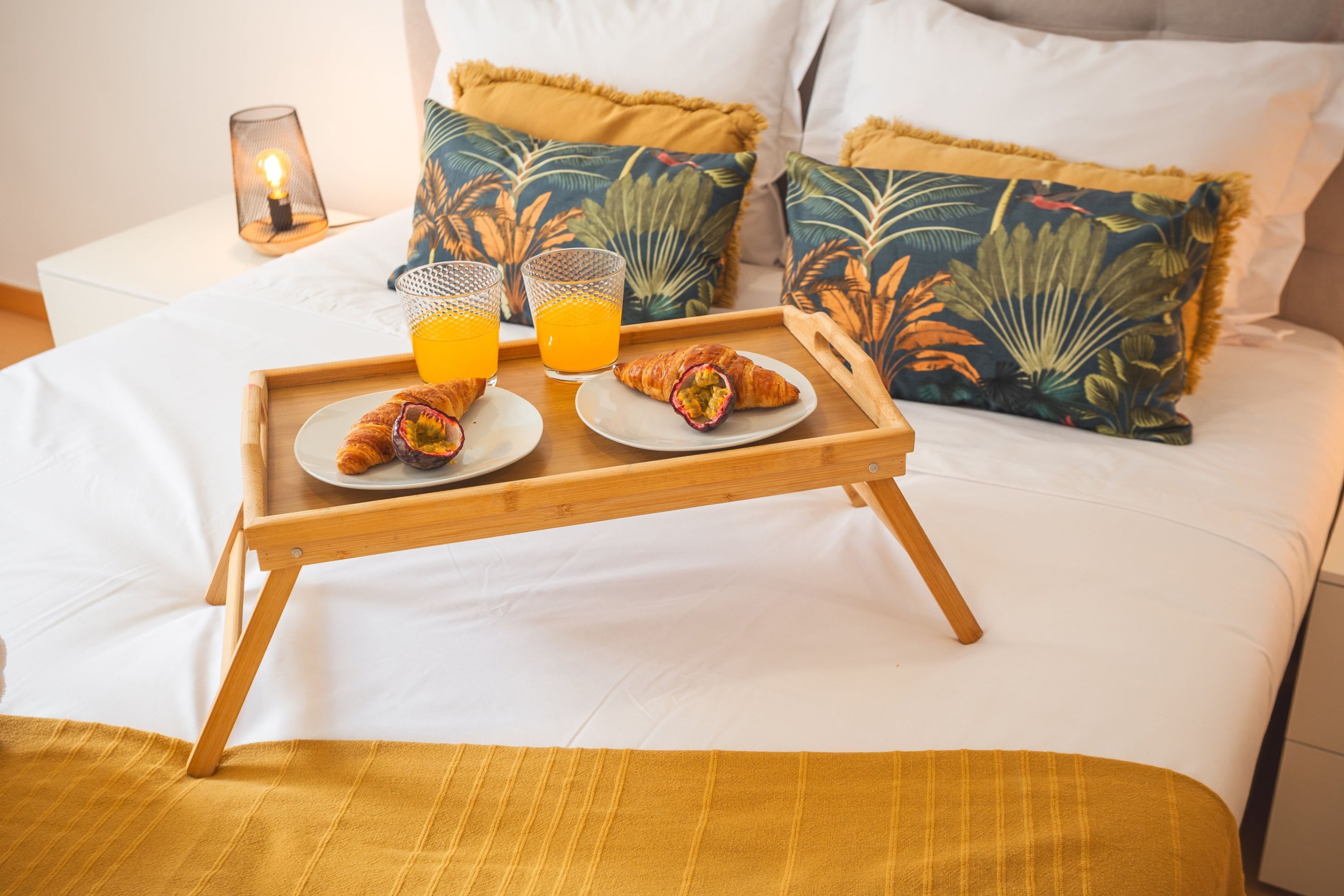 Ein Bett mit einem hölzernen Tablett, das Croissants und Orangensaft enthält, ein Nachttisch mit einer Kerze und Kissen mit tropischem Muster.