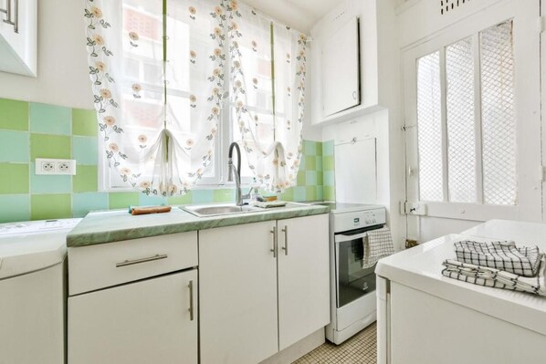 Une cuisine avec des meubles blancs, un évier, une cuisinière et une fenêtre ornée de rideaux à motifs floraux.
