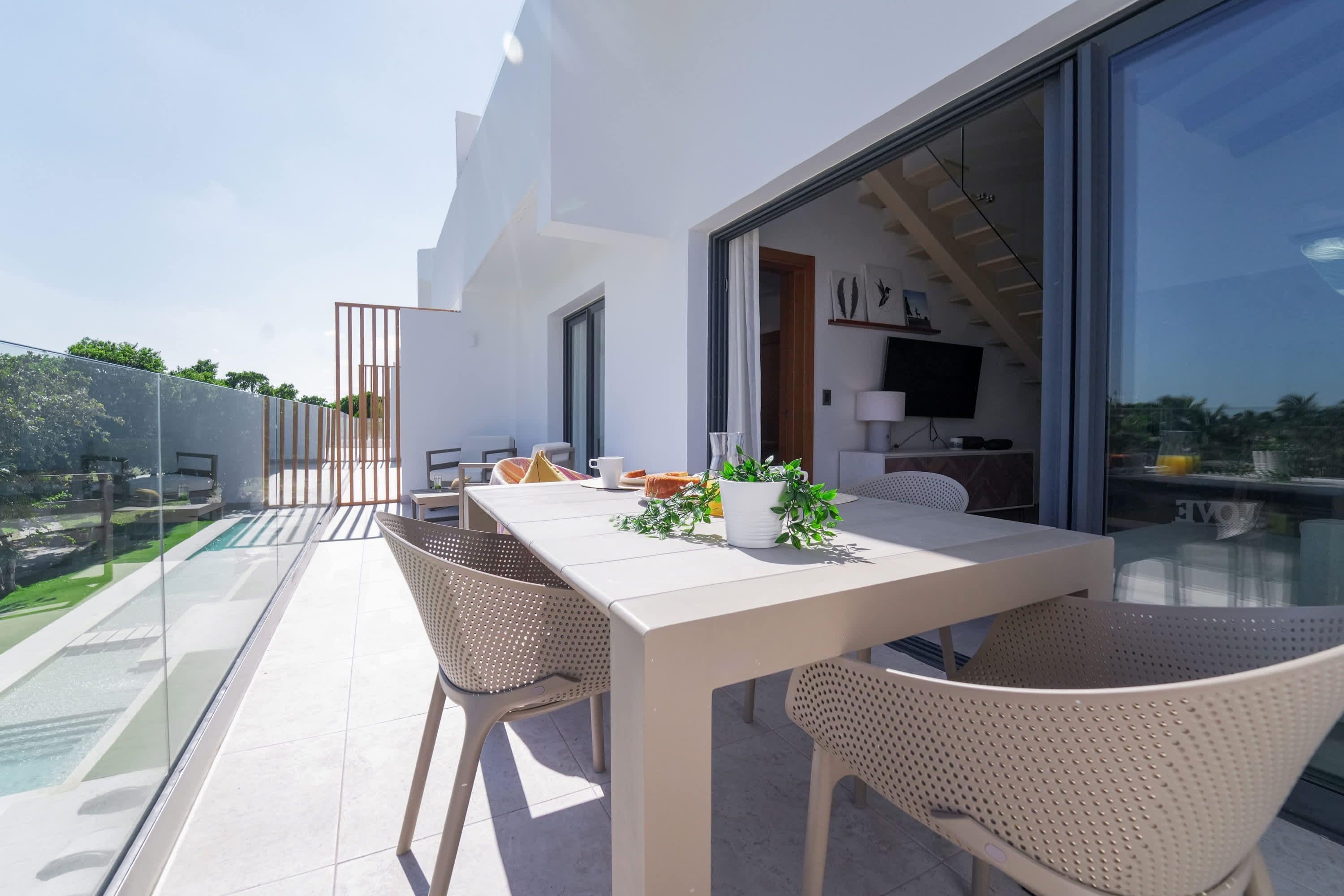 Un espacio moderno para comer al aire libre con una mesa blanca y sillas de mimbre, con vistas a una piscina y follaje.