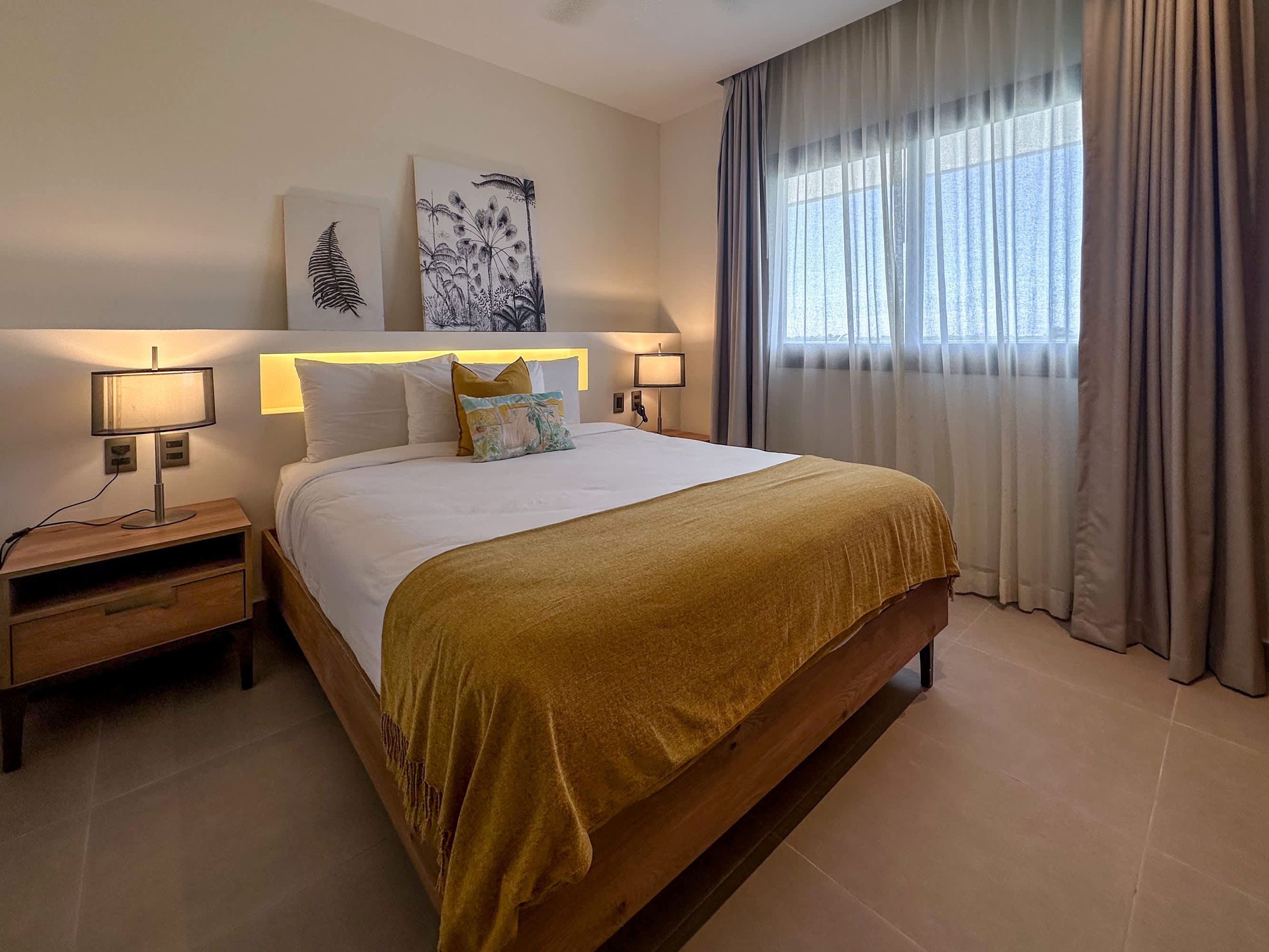 Ein ordentlich eingerichtetes Hotelzimmer mit einem großen Bett, zwei Nachttischen mit Lampen, einem Fenster mit Vorhängen und zwei Wandbildern.