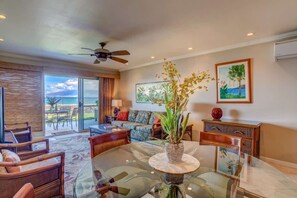 A spacious living room with a glass table, wicker furniture, a ceiling fan, and a view of the outdoors.