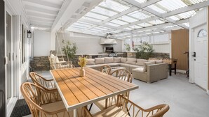 A covered patio with a wooden table and wicker chairs, a stone fireplace, and a large sofa.