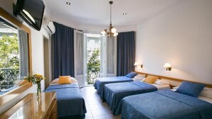 Ein Hotelzimmer mit vier Betten, einem Fernseher, einem Schreibtisch mit Blumen in einer Vase und einem Balkon mit Vorhängen.
