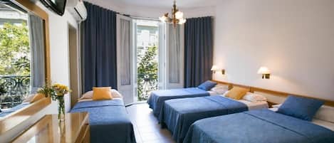 A hotel room with four beds, a TV, a desk with a vase of flowers, and a balcony with curtains.