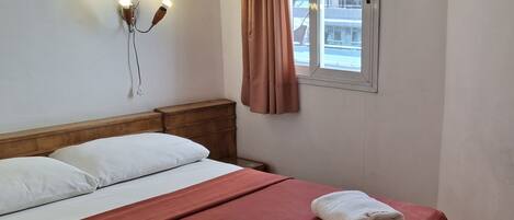 Una habitación de hotel con cama, mesita de noche, lámpara de pared, cortinas en la ventana y vistas a edificios.