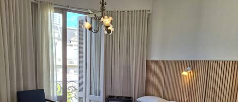 Quarto de hotel com duas camas, uma cadeira, uma cabeceira de madeira, mesa de cabeceira e varanda com cortinas.