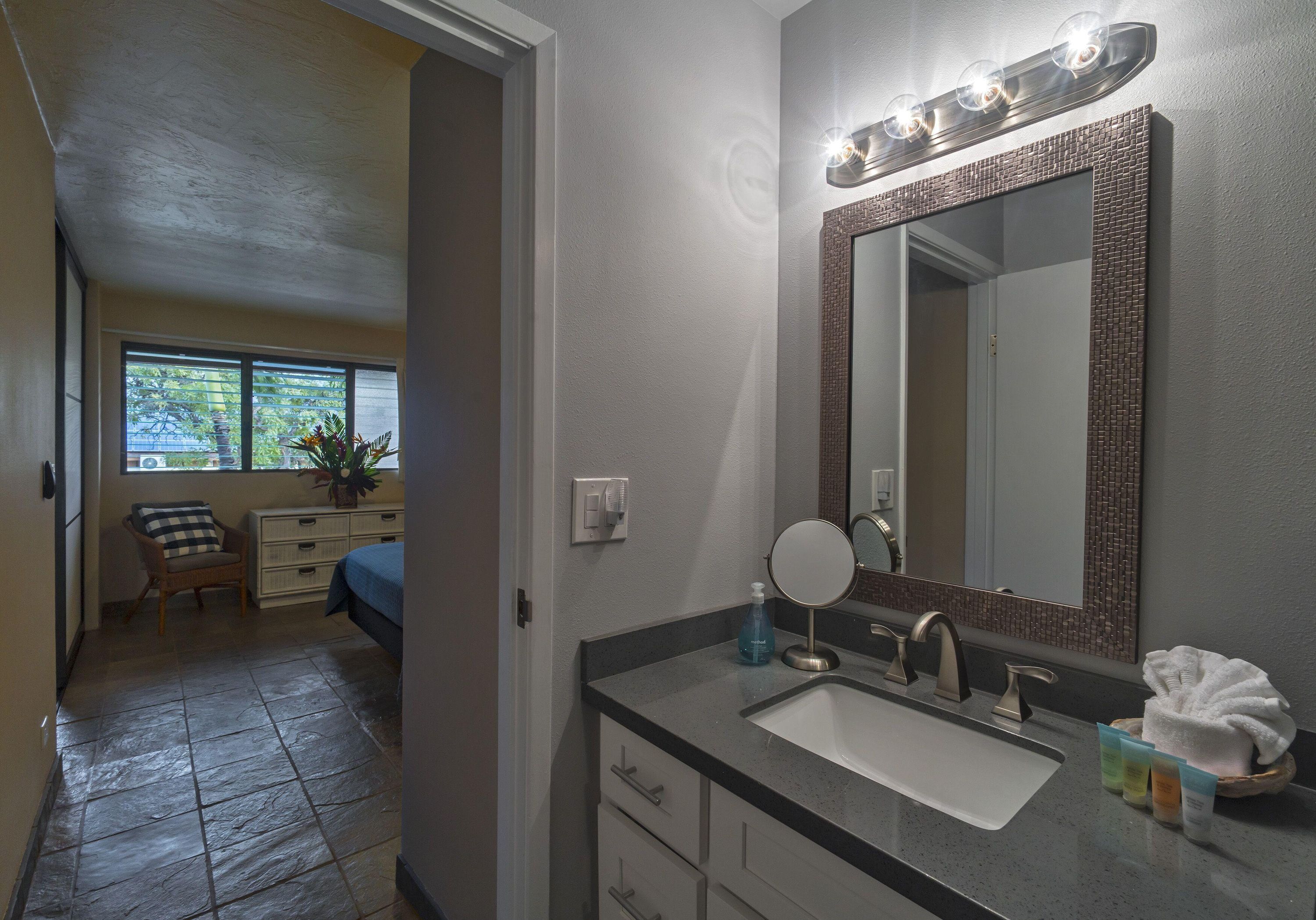 Une salle de bain avec un grand miroir, un lavabo et un meuble-vasque. Il y a une fenêtre avec des stores et une plante posée sur le rebord de la fenêtre.