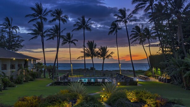 A pool area with a view of the ocean, surrounded by palm trees and well-lit pathways. - Waiohuli Beach Hale C212 (Kihei)